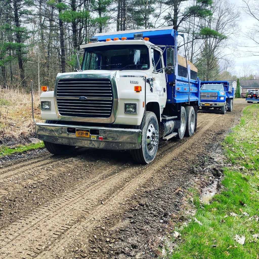 Stone Kote Construction Ford dump truck with blue dump bed on a gravel road, with trees and a second truck in the background.