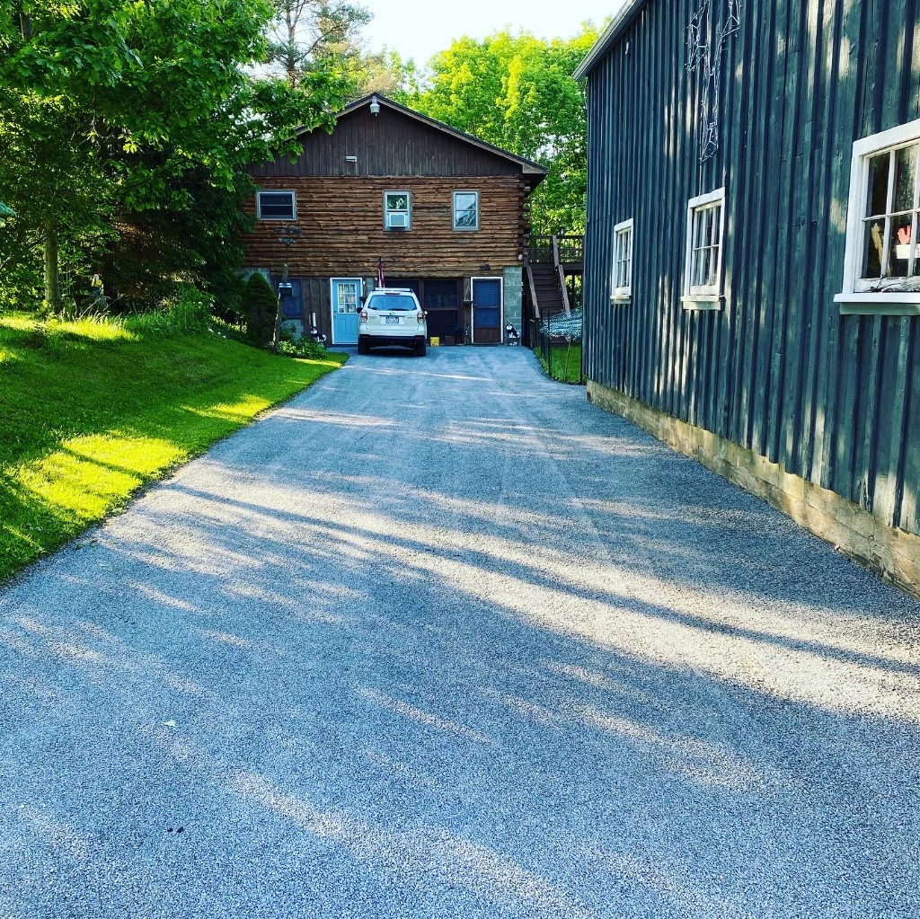Wide light grey stone driveway beside a barn and two-story home, with long afternoon shadows across the surface.