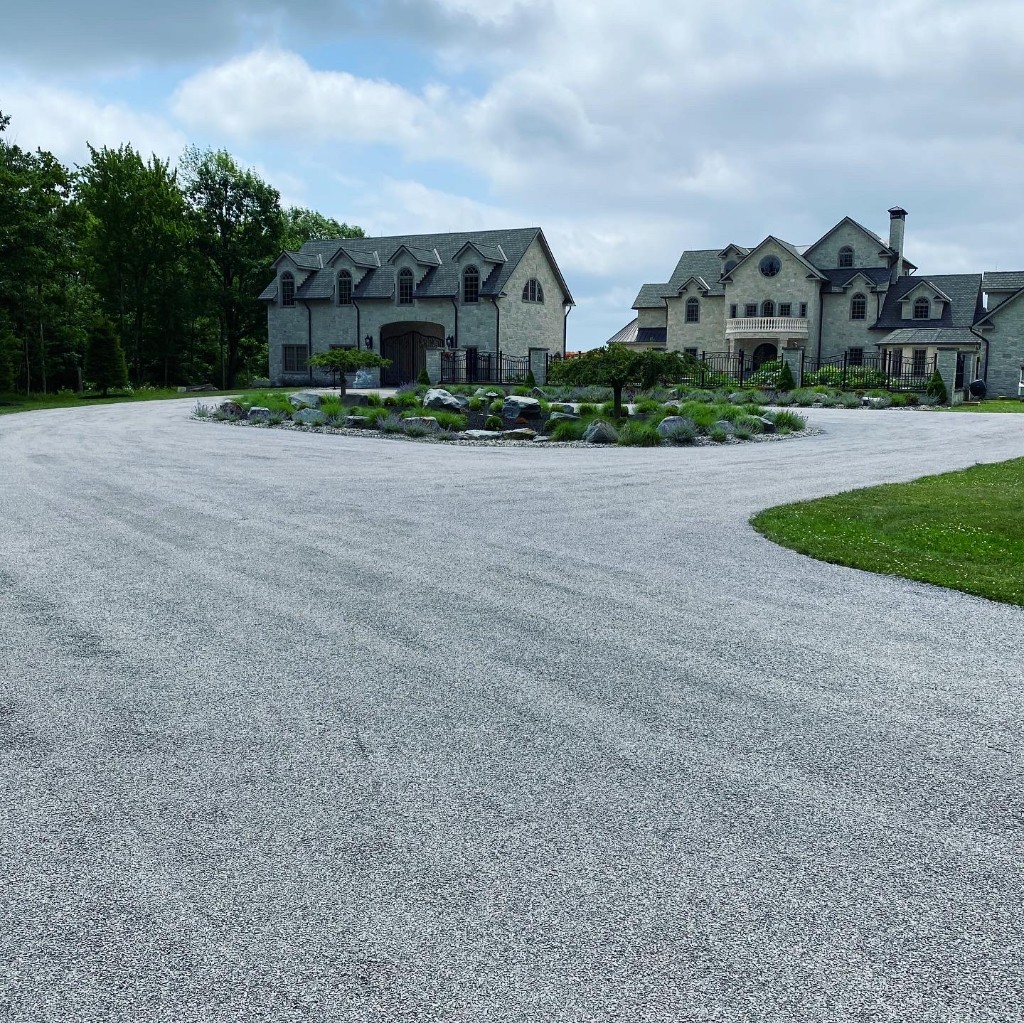 Expansive light stone driveway curving around a landscaped island with boulders and plantings in front of large stone buildings.