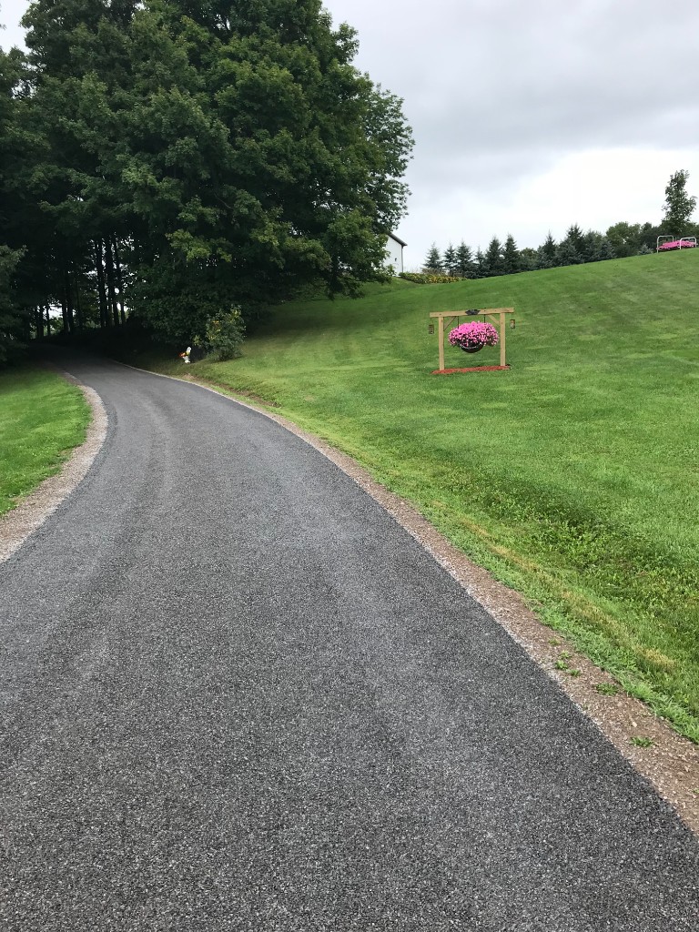 Dark grey stone and oil driveway curving uphill beside manicured lawn, trees, and a decorative flower basket.