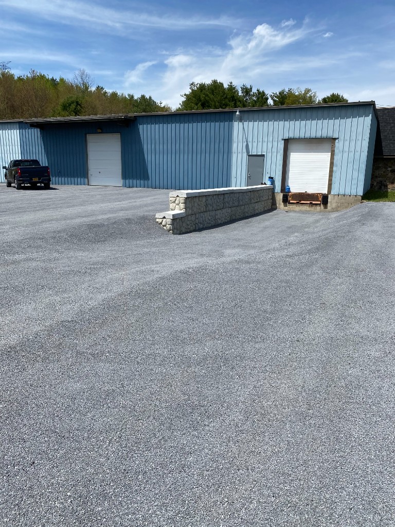 Industrial building with light blue siding and a wide grey gravel yard, low stone wall, and pickup truck for scale.