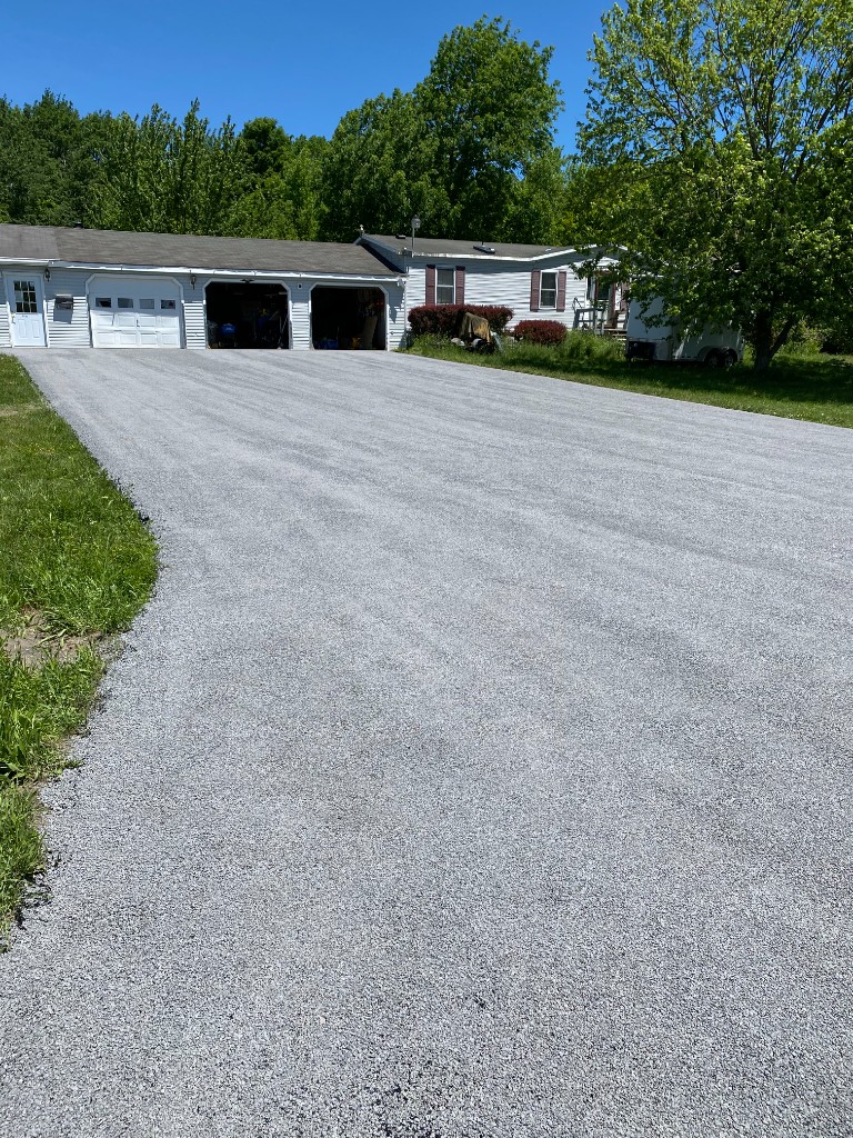 Fresh light grey stone and oil driveway leading to a white ranch home with three-car garage on a sunny day.