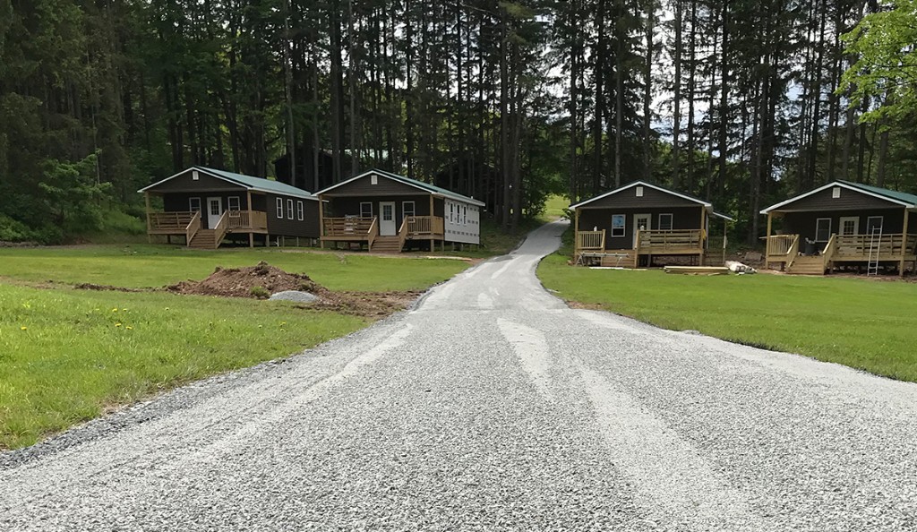 Wide grey gravel private road leading past small cabin-style buildings with green lawns and forested hillside in the background.