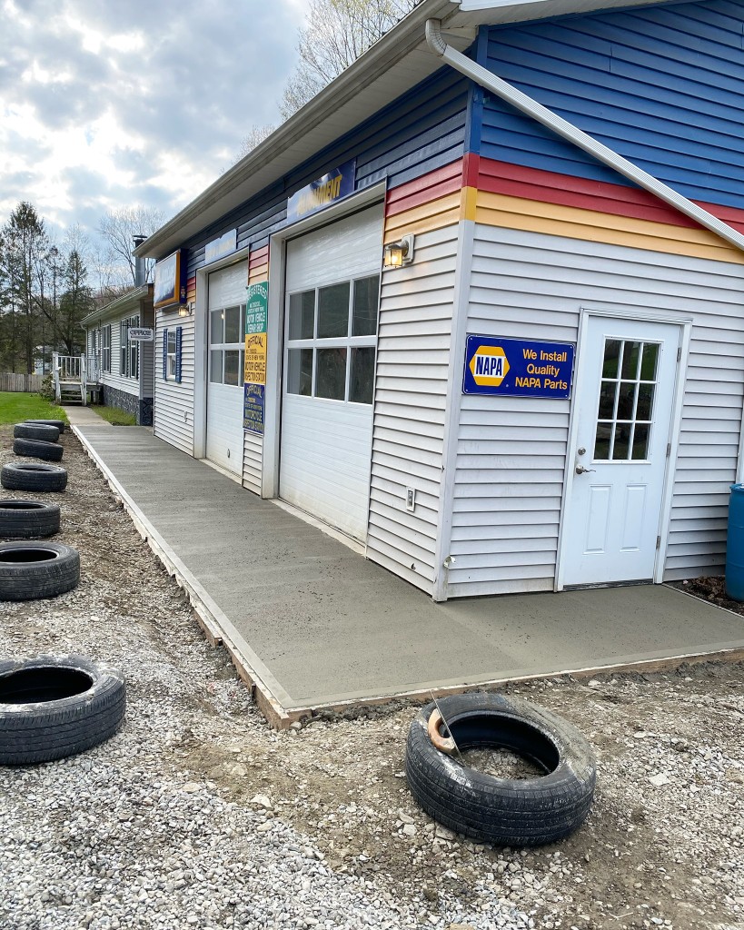 Freshly finished concrete walkway and apron in front of a commercial auto shop with garage bays, forms along the edges, and gravel surrounding the pour.