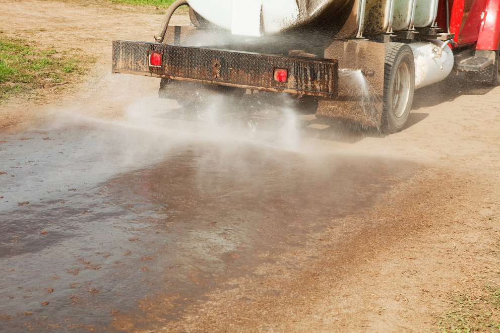 Tank truck on a gravel road spraying dust oil or dust suppressant from rear nozzles, darkening the treated strip behind the vehicle.