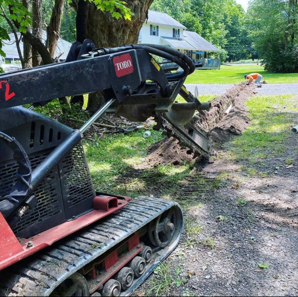 Toro compact track loader with trencher attachment cutting a straight trench through a residential lawn beside a gravel path.