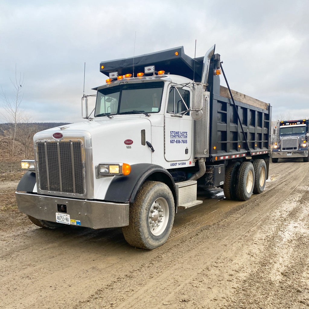 Stone Kote Construction Peterbilt tri-axle dump truck with black dump body on a muddy unpaved road at a job site.