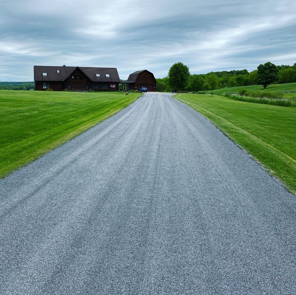 Long residential stone and oil driveway with even grey chip surface, green lawns on both sides, leading toward a log home and outbuildings under a cloudy sky.